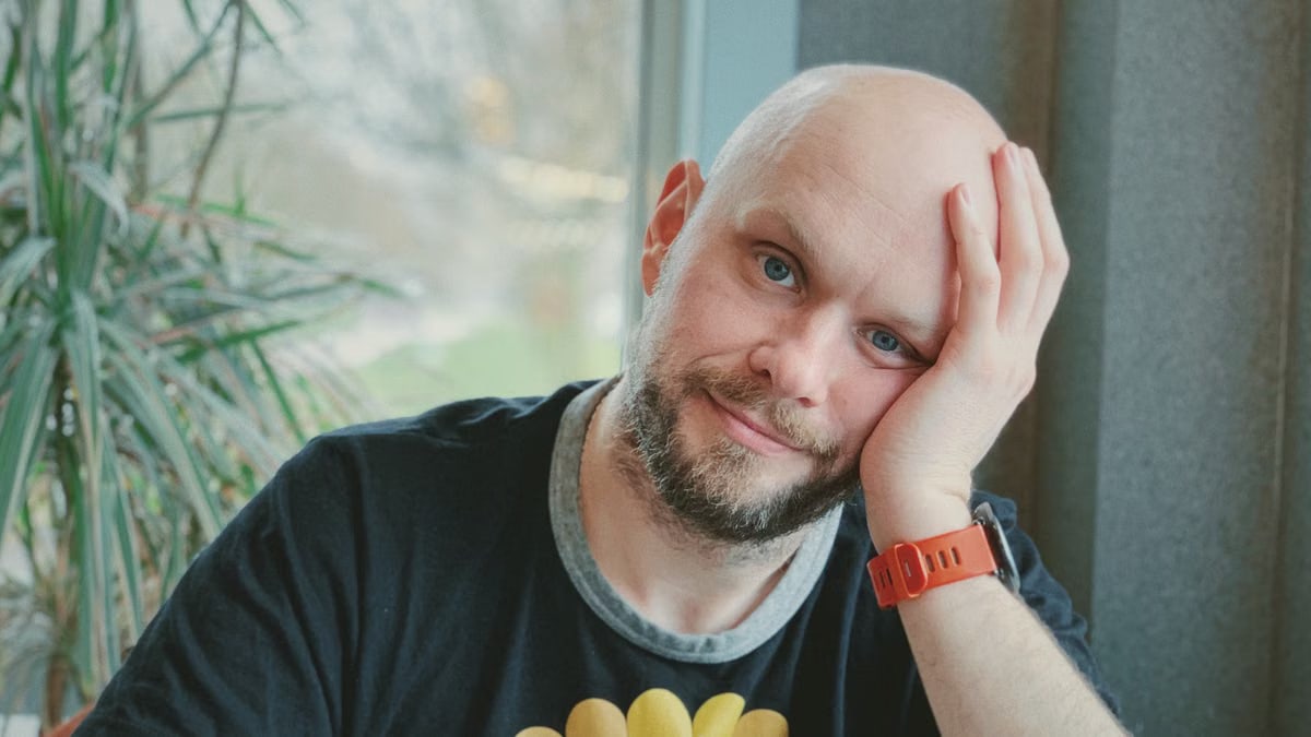 Casual portrait of a bald man sitting indoors, wearing a colorful t-shirt and red watch, with plants in the background.